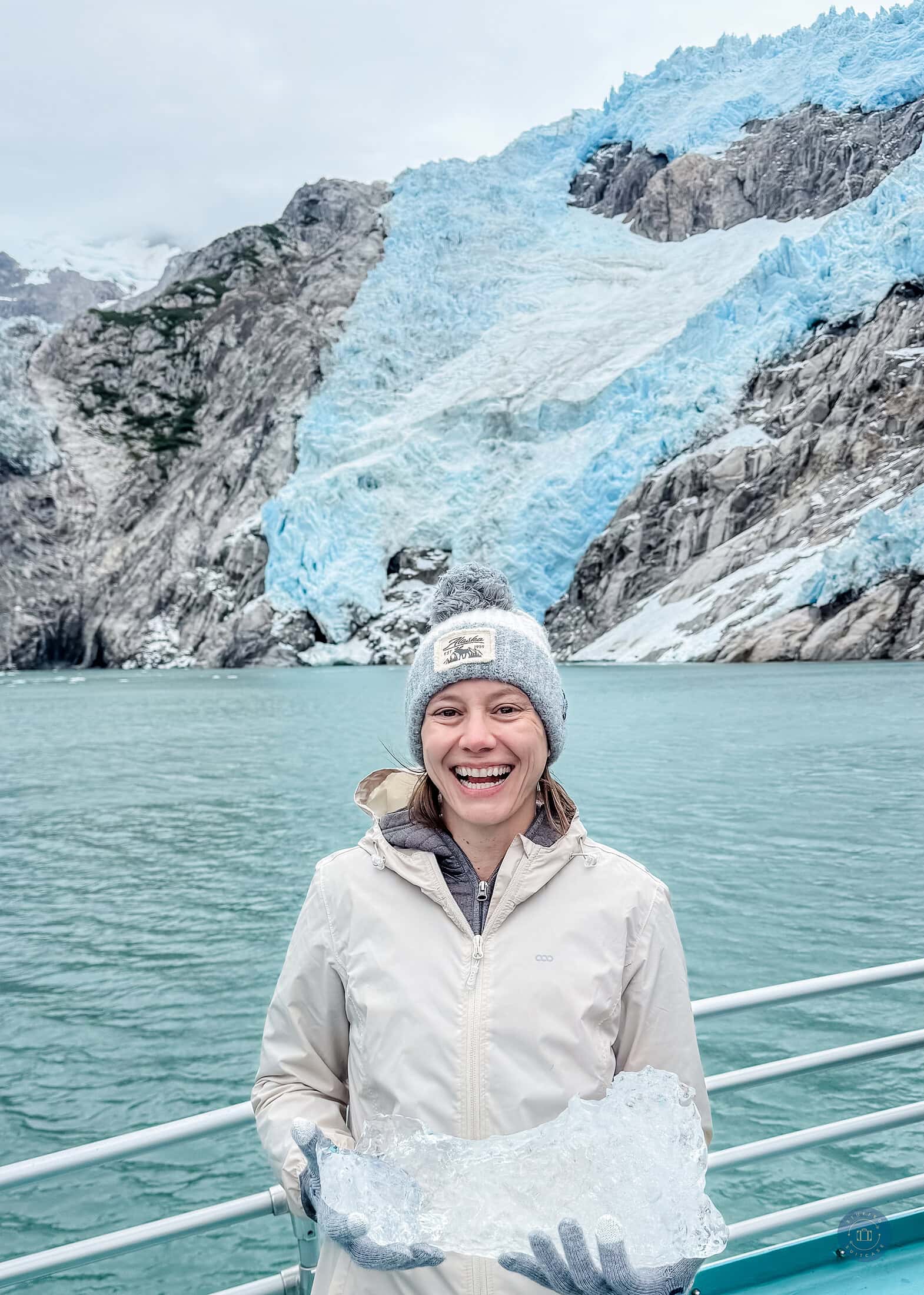 A woman is holding a piece of glacier ice in front of an Alaska Kenai Fjords glacier.