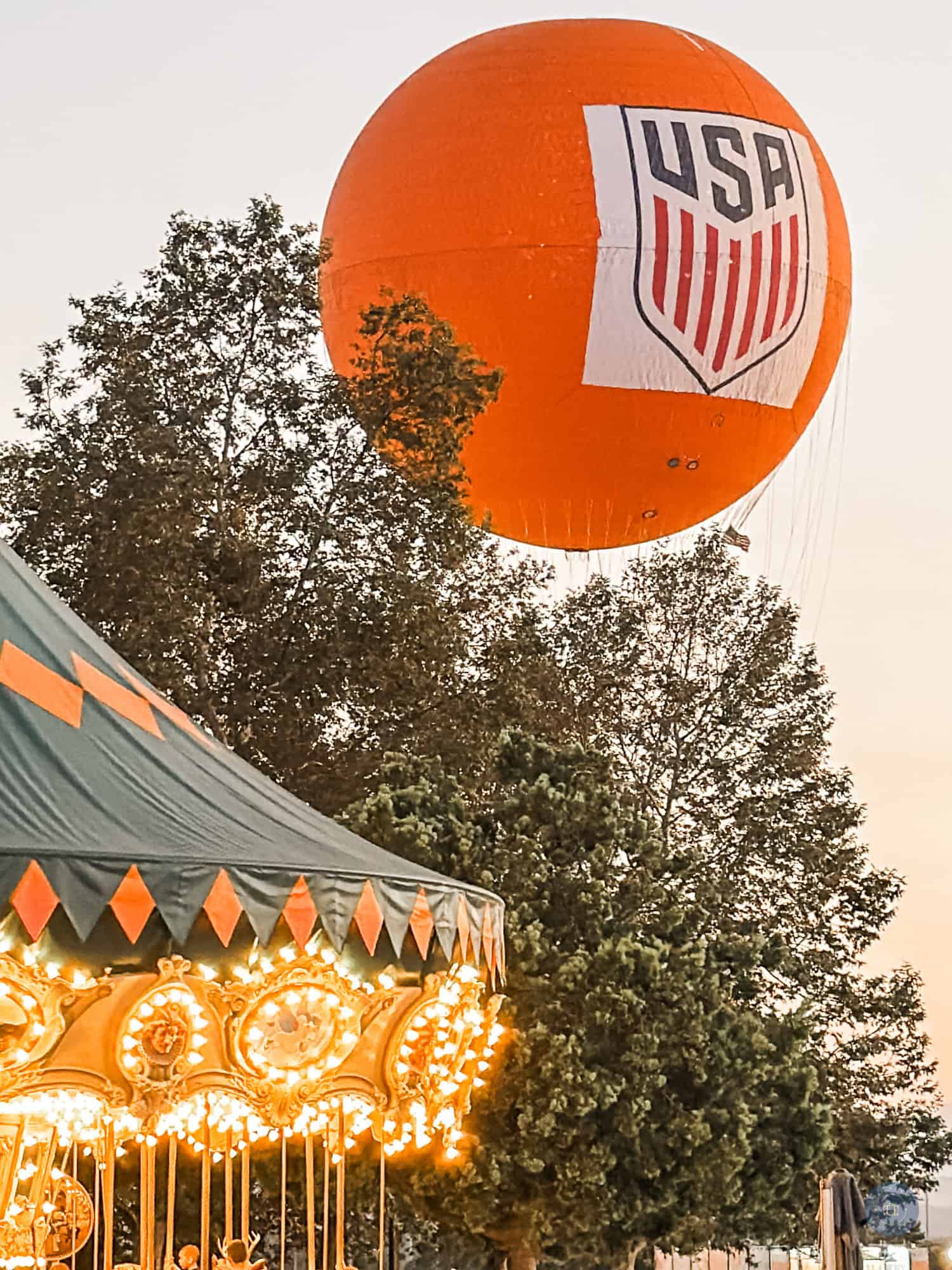 Large orange USA Great Park balloon floating above trees at Irvine California with carousel lit up in foreground.